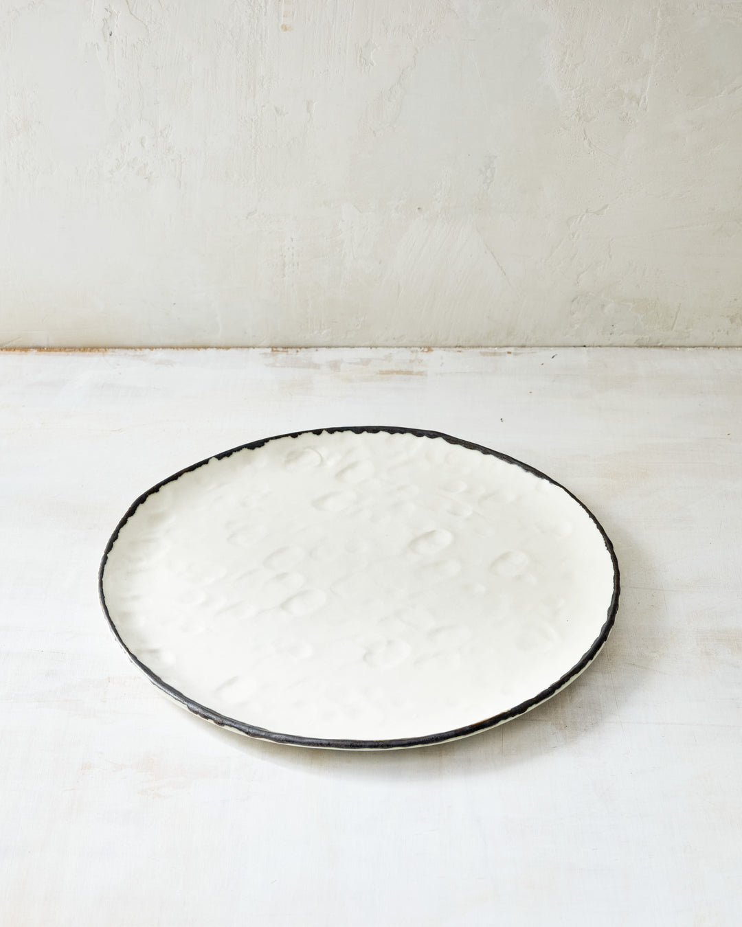DBO HOME Pinch Dinner Plate in hand-formed white porcelain with a bronze-edged rim. Subtle indentations and an organic silhouette highlight the artisanal process, styled on a pale, textural backdrop.