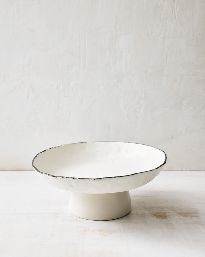 Angled view of the DBO HOME Pinch Fruit Bowl, handcrafted in creamy white porcelain with a bronze-edged rim. The elevated pedestal base and softly dimpled surface create a striking yet understated centerpiece for serving or styling.