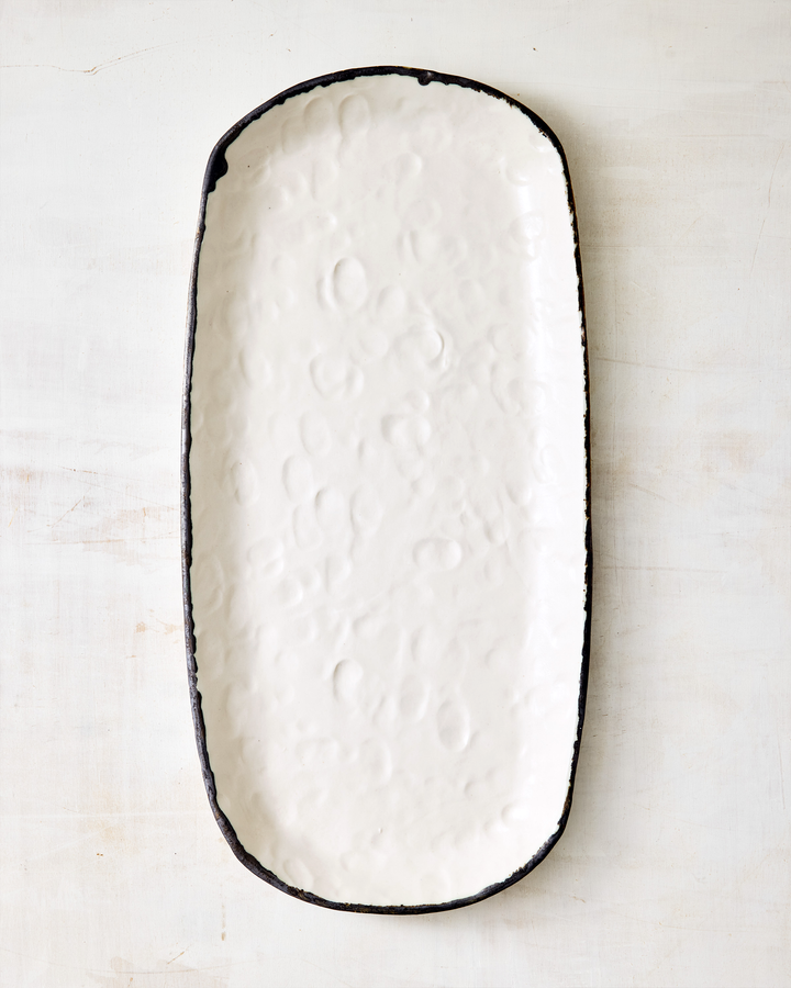 Handcrafted ceramic serving tray from DBO Home’s Pinch Collection, featuring an elongated oval shape with softly uneven edges. The creamy white glaze reveals subtle dimples and organic texture, framed by a raw dark rim. Styled on a pale, rustic surface, the tray’s minimalist elegance invites everyday use or special occasion serving.