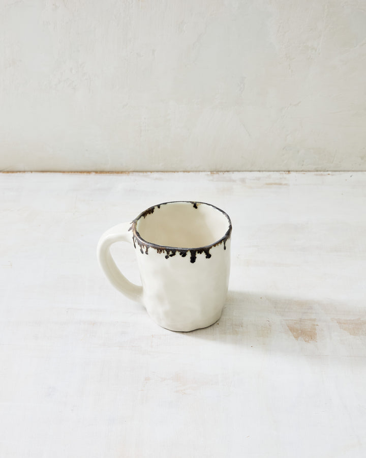 Handmade ceramic coffee mug from the DBO HOME Pinch Collection, featuring an organic pinched shape and dark drip glaze on a creamy stoneware base, perfect for coffee or tea, styled on a rustic whitewashed table.