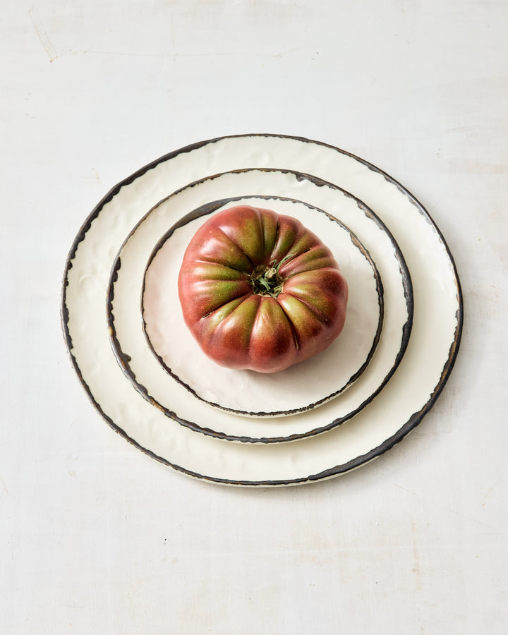 A stack of three handcrafted ceramic plates from DBO Home’s Pinch Collection, glazed in creamy white with expressive, irregular dark edges. At the center of the smallest plate sits a deep red heirloom tomato, its rippled skin adding organic charm. Set against a softly textured whitewashed background, the composition feels rustic, fresh, and artfully simple.