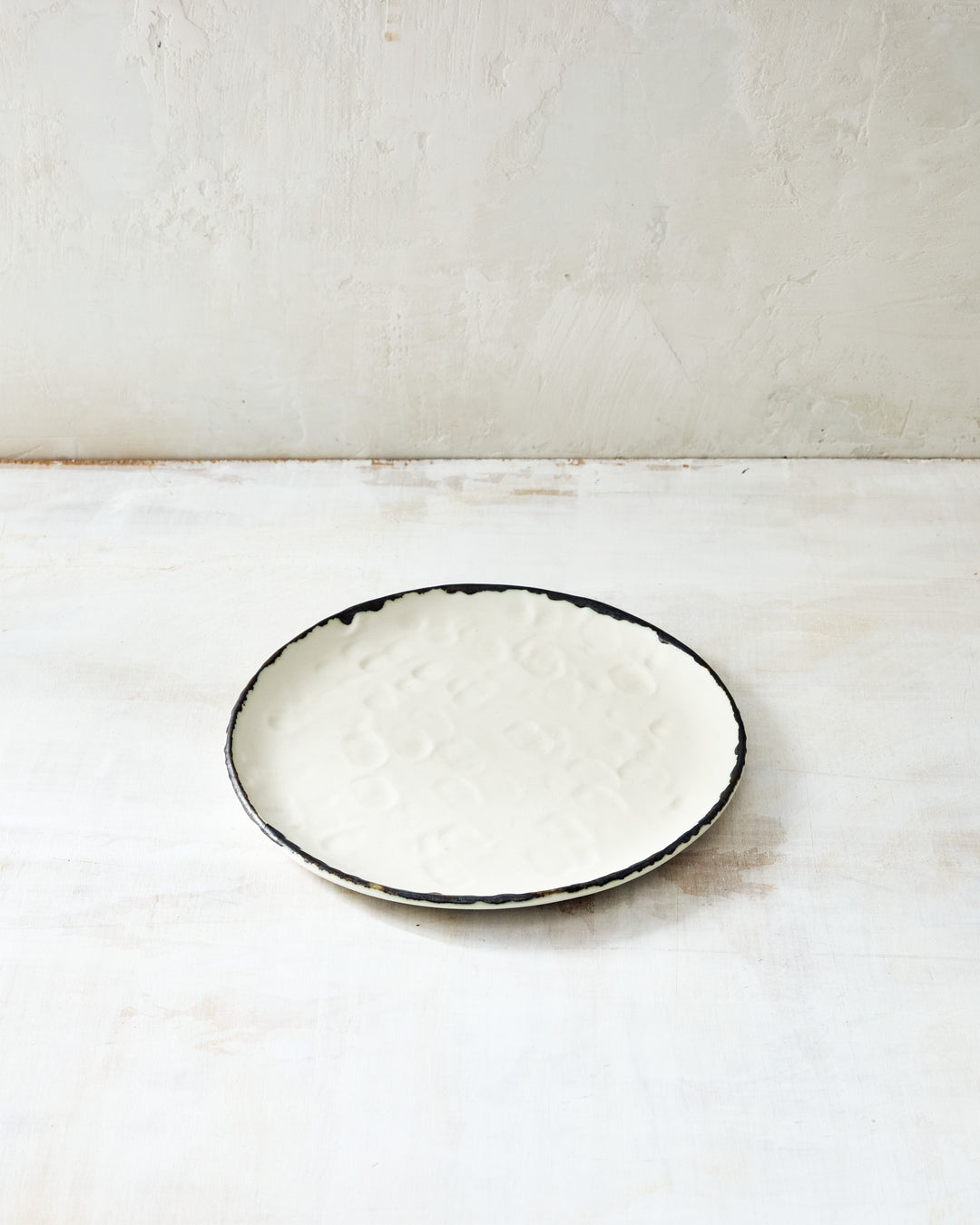 DBO Home Pinch Salad Plate in cream porcelain with bronze rim, shown from above on a textured whitewashed surface. The hand-formed plate features subtle fingerprint-like impressions, highlighting its artisanal craftsmanship.