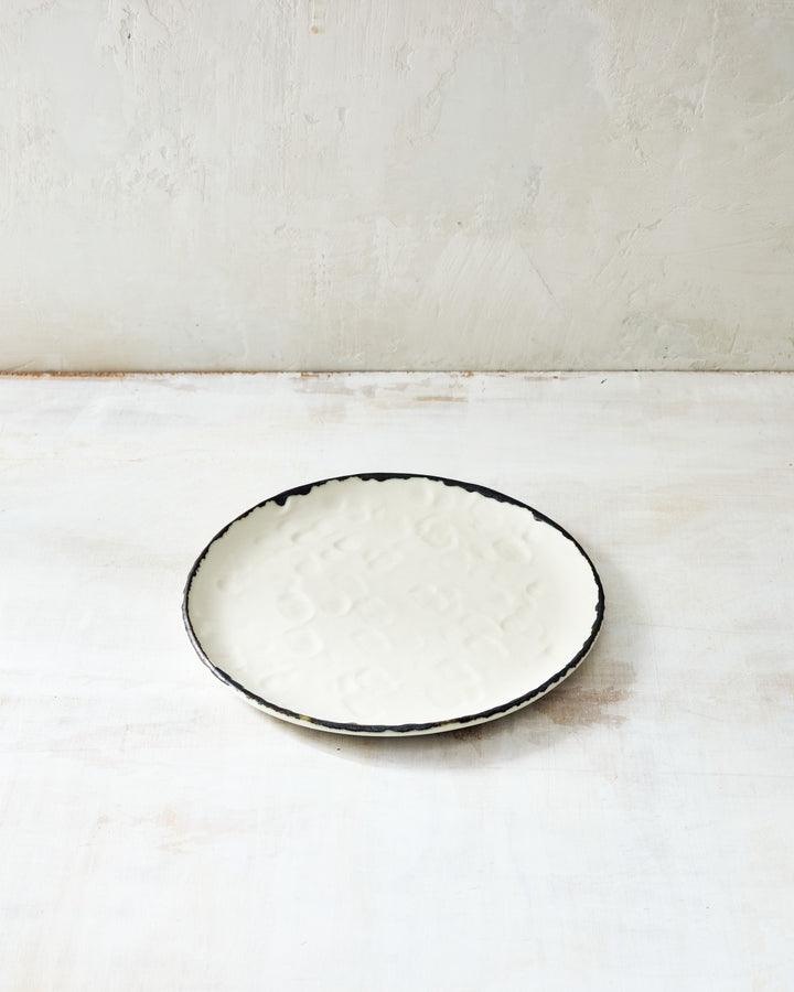 DBO Home Pinch Salad Plate in cream porcelain with bronze rim, shown from above on a textured whitewashed surface. The hand-formed plate features subtle fingerprint-like impressions, highlighting its artisanal craftsmanship.