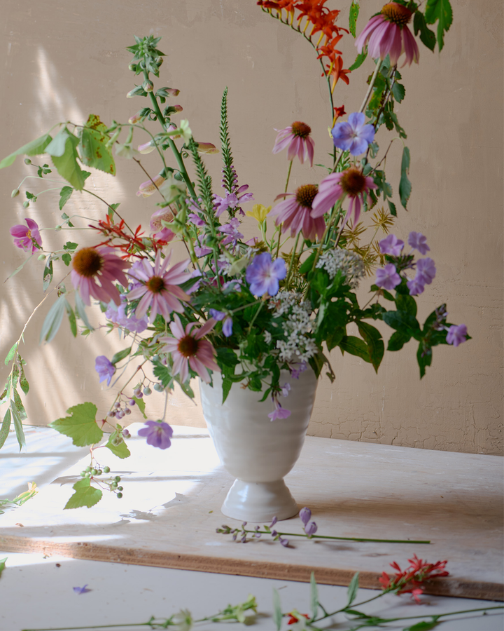 DBO HOME Bare Pedestal Vase in Snowflake porcelain, filled with a wild, seasonal arrangement by Romane Recalde of Le Jardin Flower Farm. Photographed by John Gruen in soft natural light, the vase’s smooth form anchors the airy, organic composition. [Styling: Romane Recalde; Photography: John Gruen]
