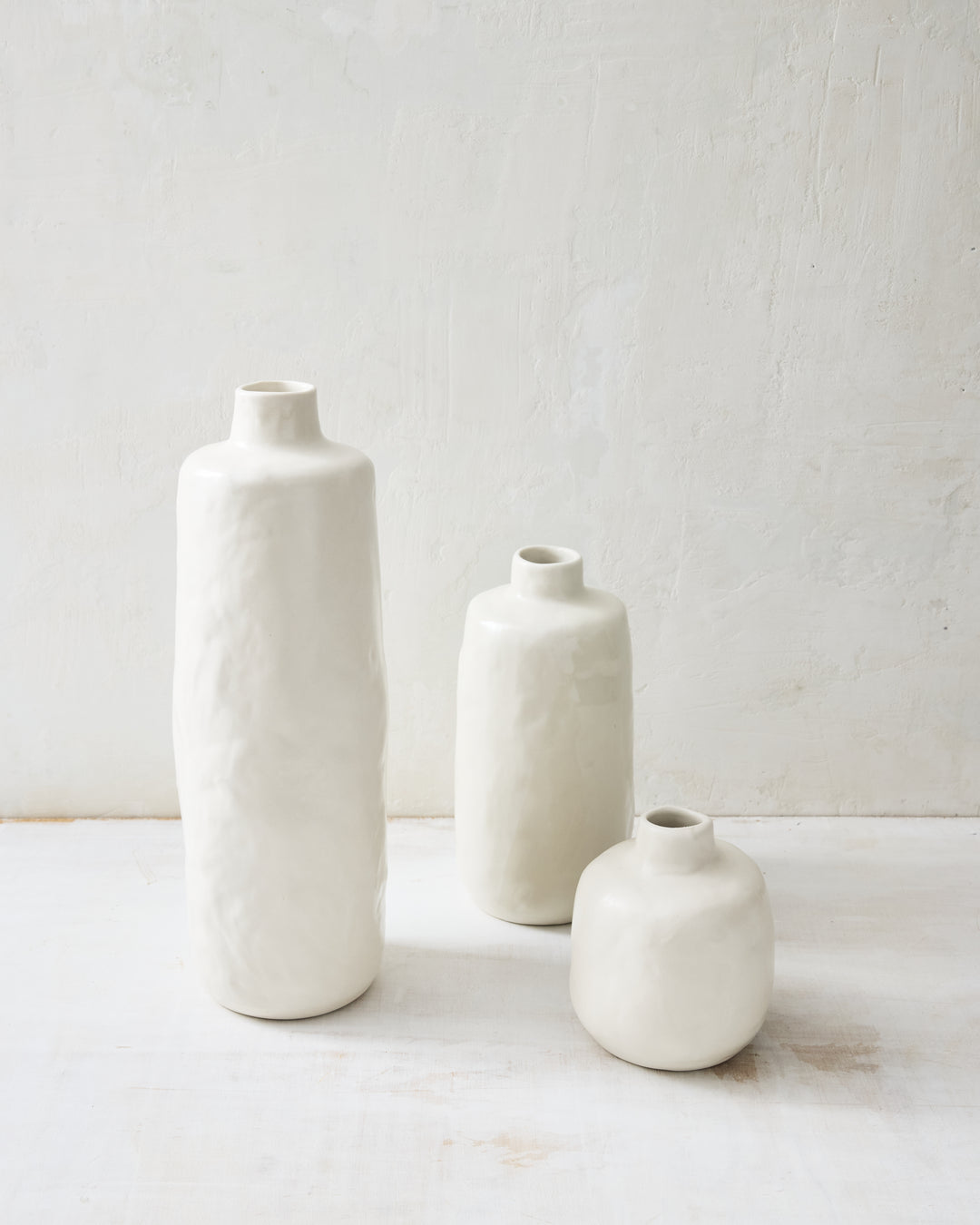 Group of handmade Wabi ceramic vases in soft Foam glaze, featuring organic, irregular forms and a matte white finish, arranged on a white surface against a textured plaster wall.