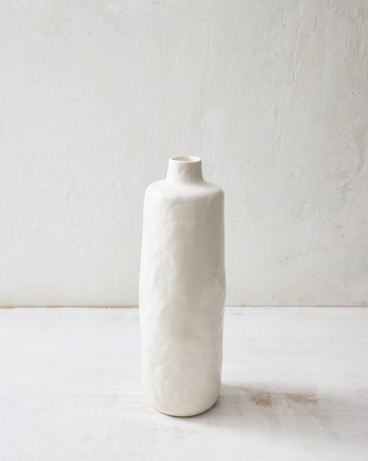 Tall Wabi ceramic vase in Foam glaze with an elongated, sculptural profile, hand-formed texture, and foam white finish, styled against a softly textured plaster backdrop.