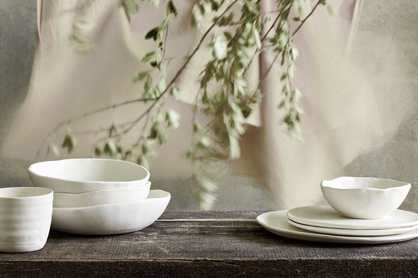 DBO HOME artisan ceramic minimalist white porcelain plates and bowls stacked on a rustic dark wood table in front of a soft natural cloth background with blurred leafy branches dipping down