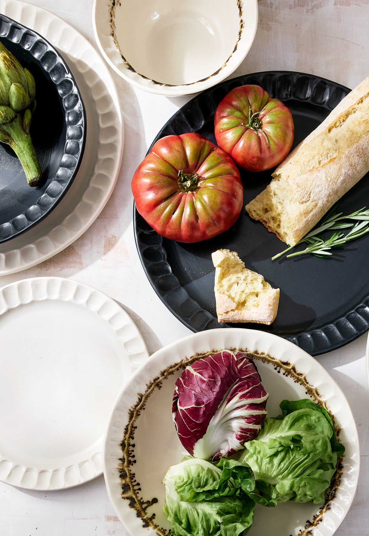 Overhead view of the DBO HOME Nonna dinnerware collection, including dinner plates, salad plates, small bowls, and pasta bowls in Matte Black, Snowflake, and Bronze finishes. Styled with heirloom tomatoes, fresh greens, and rustic bread on a whitewashed surface, highlighting the handmade textures and inviting forms.