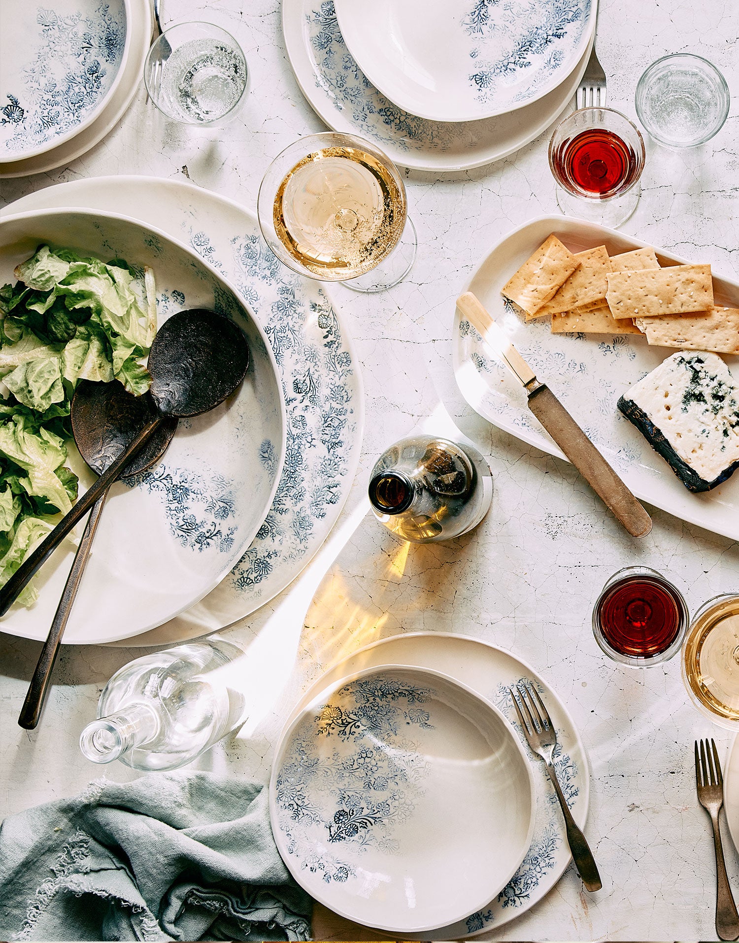 An elaborate and elegant sunny summer table setting featuring white porcelain plates and serving pieces partially covered by indigo Indian woodblock prints, among glasses of summer wine with salad, cheeses and crackers strewn throughout