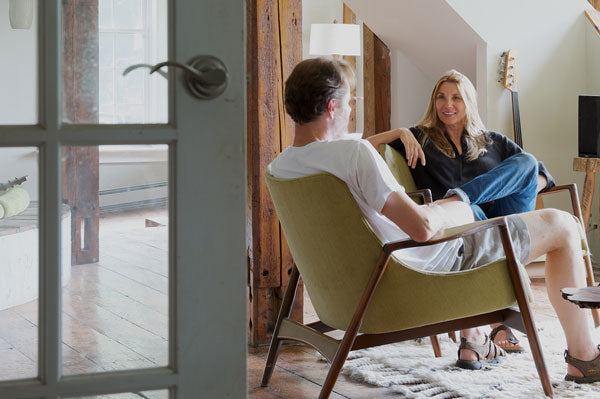 Daniel Oates and Dana Brandwein of DBO HOME sit in mid century modern green upholstered chairs in a light-filled living room with a guitar in background and a glass paneled door in the left foreground