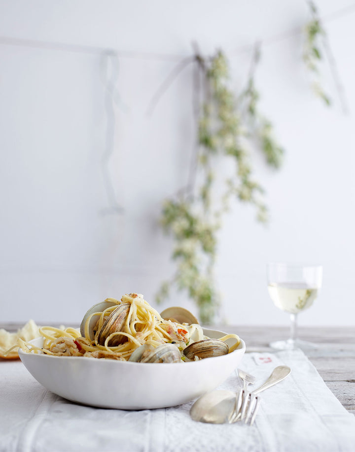 DBO HOME Artisan ceramic Bare Everything Bowl filled with clam pasta on a wooden table with vintage silverware and a glass of white wine