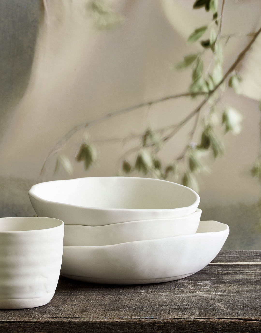 DBO HOME stack of handmade porcelain cream colored Bare Noodle Bowls and Bare Everything Bowl with artisan ceramic tumbler on natural wood table with softs linens and plant branches in the background