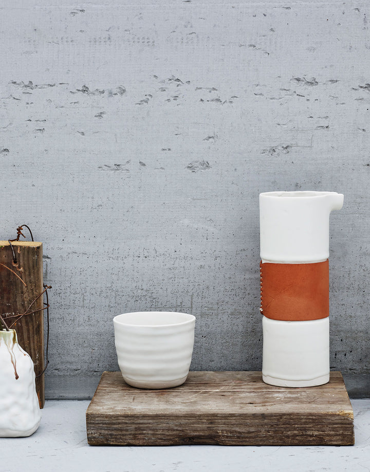 DBO HOME bare tumbler ceramic cup in white glaze with remo pitcher with leather details on a wood board against cement wall
