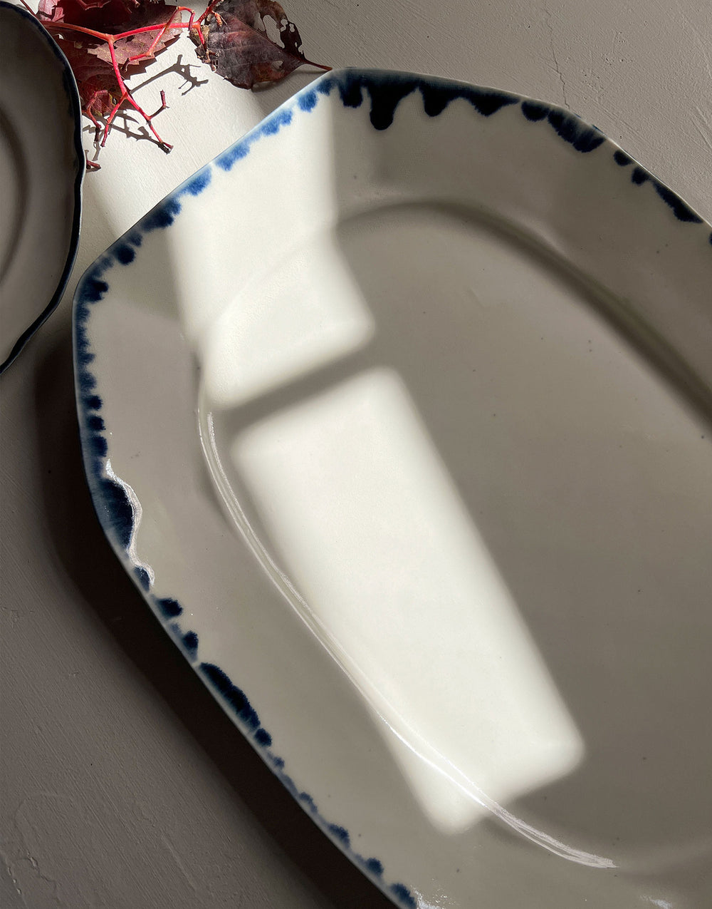 Close-up of the DBO Home Formale serving platter in Snowflake with Indigo glaze. Handcrafted ceramic with painterly cobalt rim on a soft white base, shown in dappled natural light to highlight its sculptural form and artisanal finish.