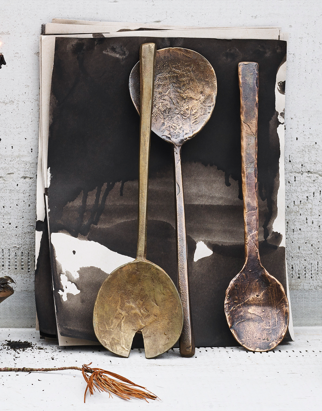 Hand cast bronze serving utensils on black ink paper with bronze spoon and pine needles on concrete background