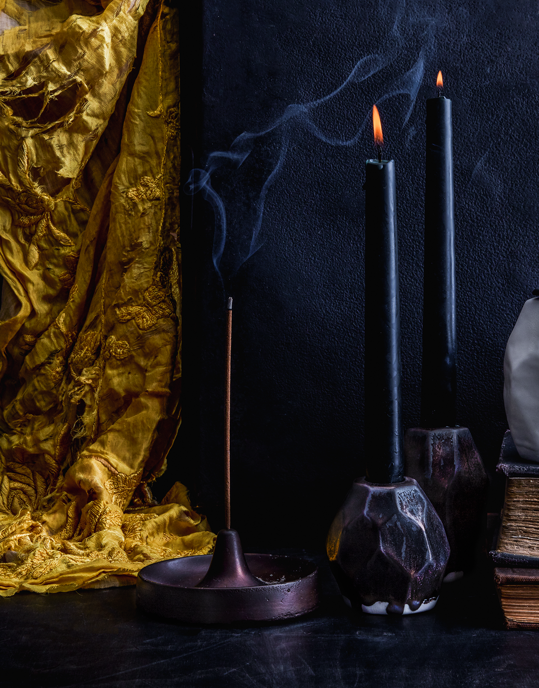 Hand-cast bronze incense burner with smokey incense, black candles, and gold fabric in a dark photography scene